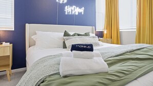 Een net opgemaakt bed met een wit hoofdbord, kussens en een opgevouwen handdoek met 'Makeup' erop. Op de muur hangt een neonbord met de tekst 'Downtown Nights'.