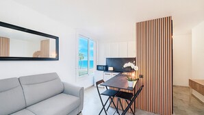 En moderne stue med en grå sofa, et spisebord med stole, et køkkenområde og et stort vindue med havudsigt.