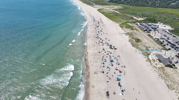 Luchtbeeld van een strand met mensen, parasols en geparkeerde auto's.
