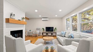 Een moderne woonkamer met een open haard, comfortabele zithoek, een televisie en een groot raam met uitzicht op bomen.