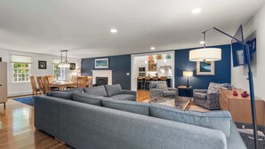 A modern living room with a large grey sectional sofa, a dining area with wooden chairs, a kitchen in the background, and a blue accent wall.
