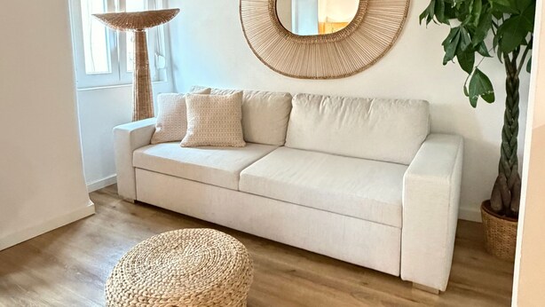 A modern living room with a light-colored sofa, a round woven ottoman, a large mirror, and a potted plant. - Arroios Elegance Residence - Lisbon Center (Lisboa)