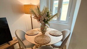 A dining area with a round table, four chairs, a vase with flowers, a lamp, and a window with blinds. - Arroios Elegance Residence - Lisbon Center (Lisboa)