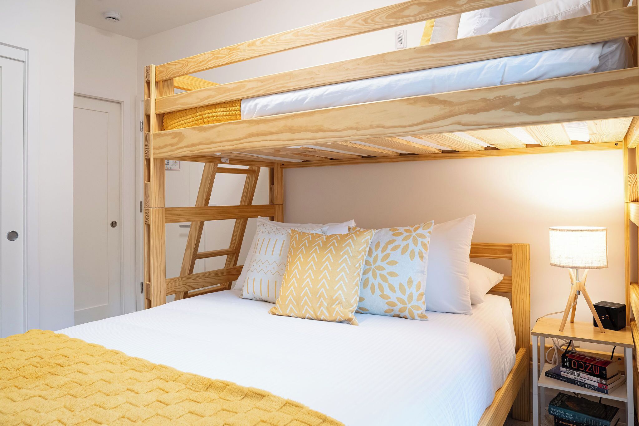 Una cama litera con estructura de madera, ropa de cama blanca y almohadas estampadas. Hay una mesita de noche con una lámpara y libros.