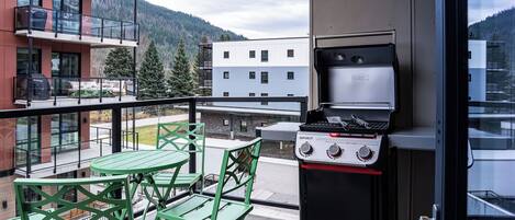 Un balcón con una parrilla, sillas verdes y vistas a edificios y montañas.
