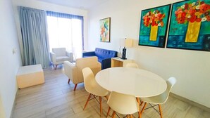 Un salon moderne avec une table ronde, des fauteuils beiges, un canapé bleu et une grande fenêtre avec des rideaux.