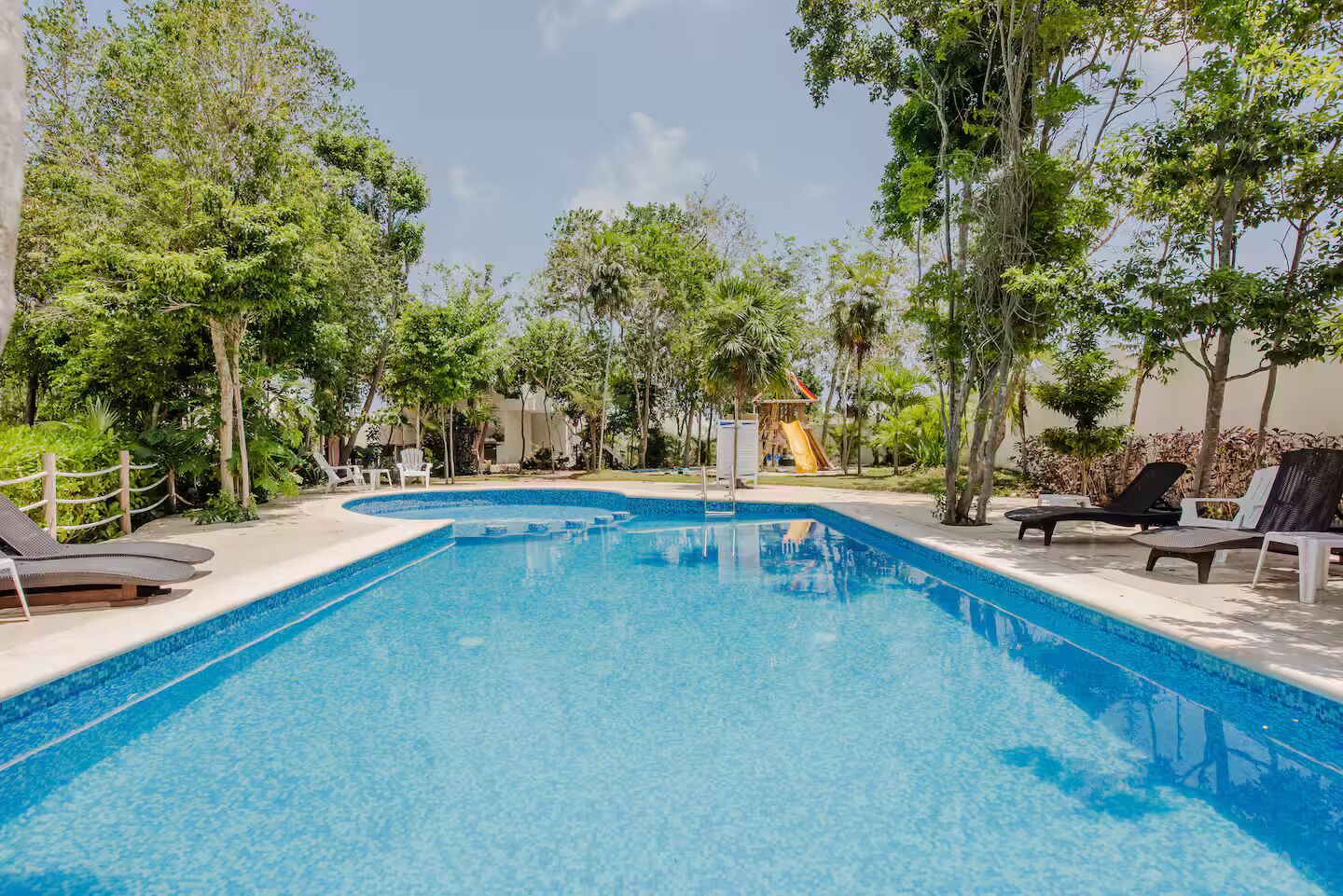 A clear blue swimming pool surrounded by lounge chairs and trees, with a slide in the background.