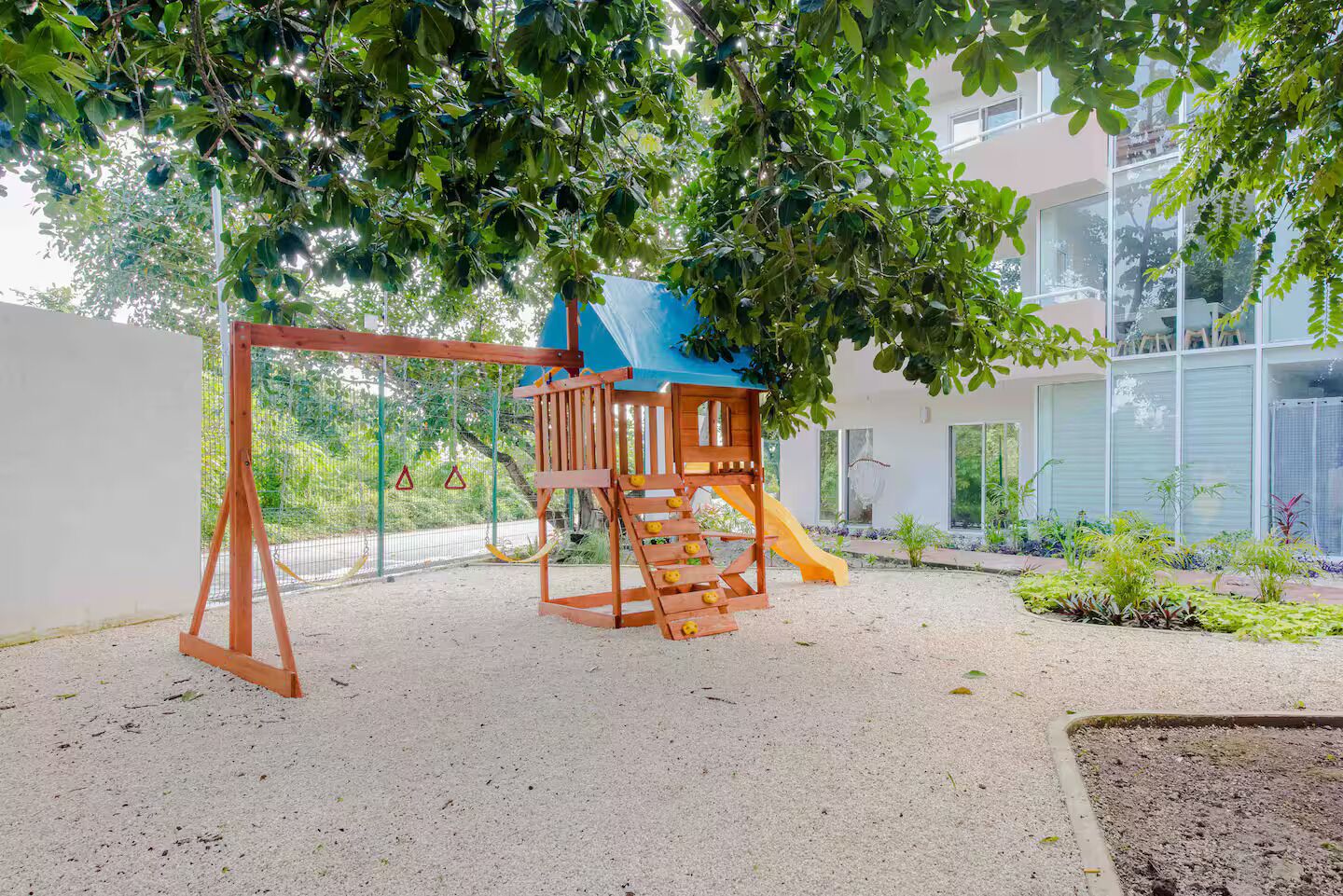 A playground with a slide and climbing structure, surrounded by a building and greenery.