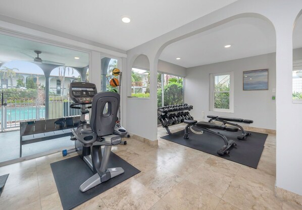 A gym with exercise equipment, a treadmill, dumbbells, and a view of a pool. - Tampa Comfort Loft on Kennedy (Tampa)