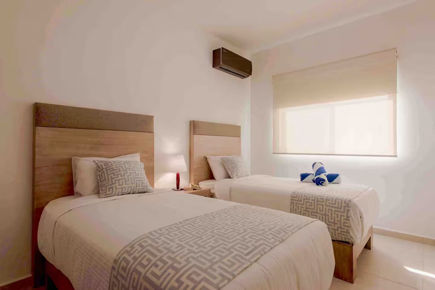 A hotel room with two beds, a window with blinds, a wall-mounted air conditioner, and a bedside table with a lamp.