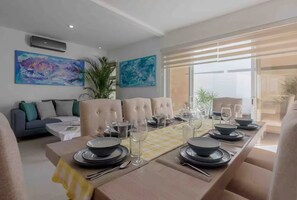 A dining area with a long table set for four, surrounded by cushioned chairs. There are two large abstract paintings on the walls, a potted plant, and a view through the window. - Premium homes Puerto Morelos (Puerto Morelos)