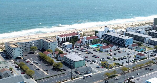 Vue aérienne d’un complexe hôtelier situé sur la côte, avec plusieurs bâtiments, un parking et une plage en arrière-plan.