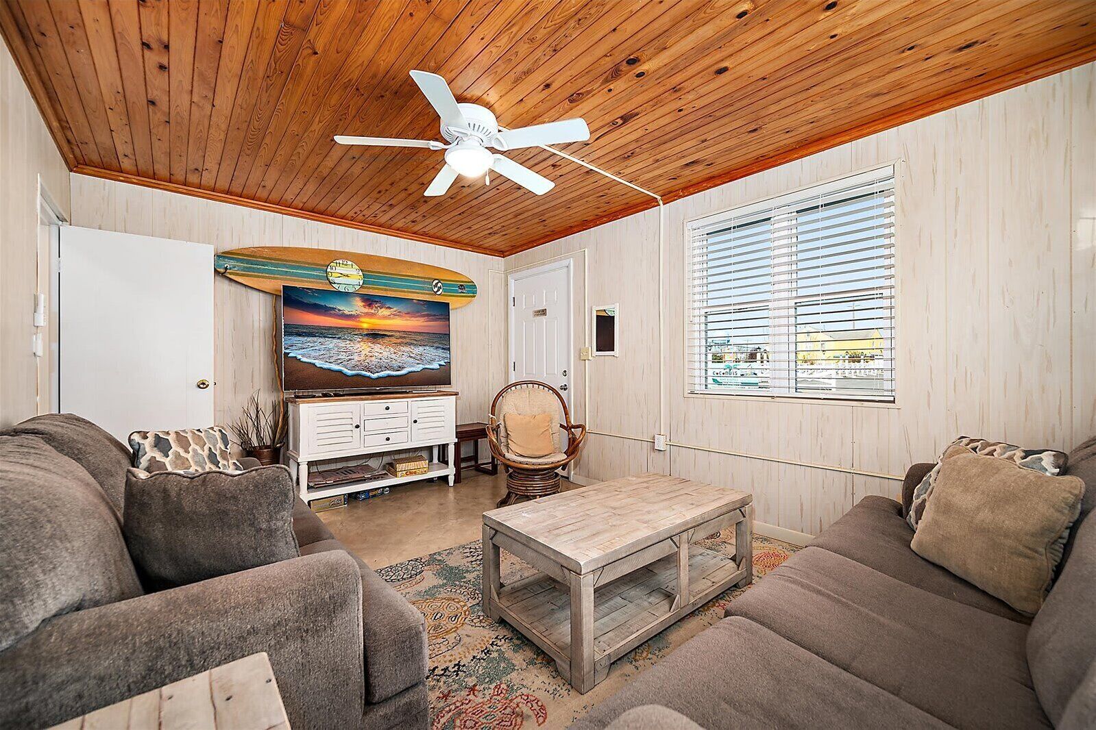 A cozy living room with a ceiling fan, a large flat-screen TV, a wooden coffee table, and a comfortable sofa set.