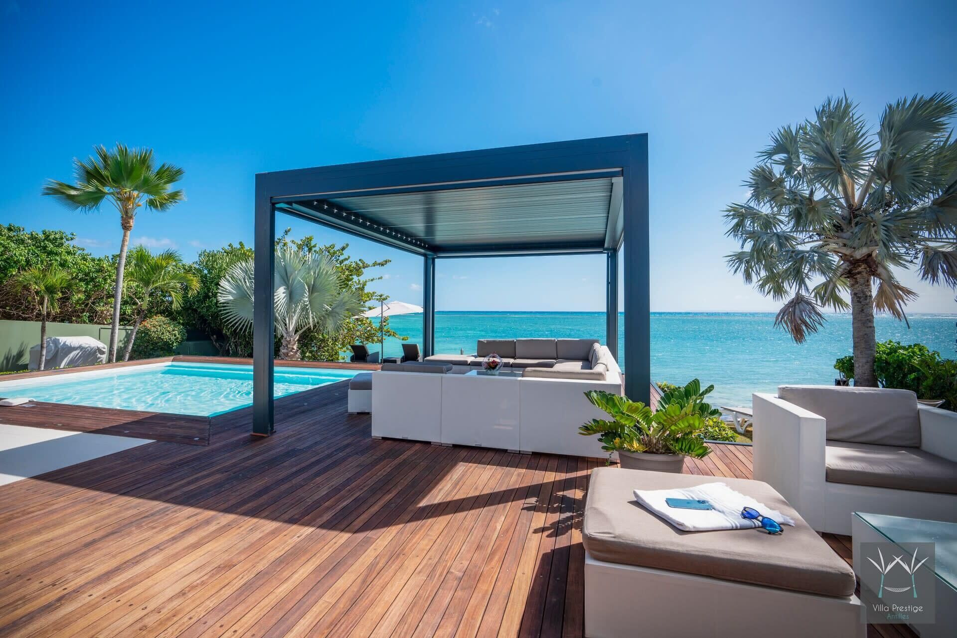 Un área de piscina exterior moderna con cubierta de madera, muebles de descanso blancos y una zona de estar cubierta con vista al océano.