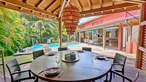 A wooden deck with a round table set for dining, overlooking a swimming pool and lush greenery. - Coco Verde (Petit-Bourg)