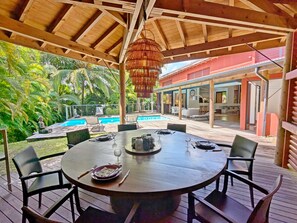 Eine Holzterrasse mit einem runden Esstisch, mit Blick auf einen Swimmingpool und üppiges Grün.