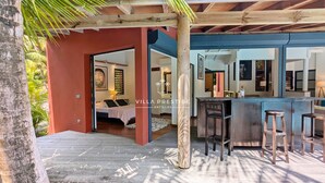 A villa with a wooden deck, a bar area with stools, and a bedroom with a bed and a nightstand. - Coco Verde (Petit-Bourg)