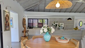 A dining area with a round wooden table, wicker chairs, a vase of white flowers, and a hanging light fixture. - Rubis |Exclusivité (Saint François)