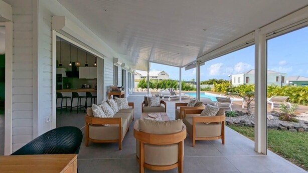 Een overdekt terras met meerdere stoelen en een tafel, een keuken met bar zitplaatsen, en uitzicht op een zwembad en groen.