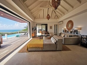 A spacious living room with a large sofa, a coffee table, and a view of the ocean through a sliding glass door. - Villa Ô | Elegant villa with sea view and walking distance to the beach (Saint-François)