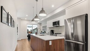 Une cuisine moderne dotée d’un îlot central, d’appareils électroménagers en acier inoxydable, de luminaires suspendus et offrant une vue sur l’espace de vie.
