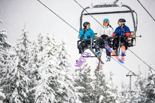 Three people on a ski lift with skis and poles, surrounded by snow-covered trees.