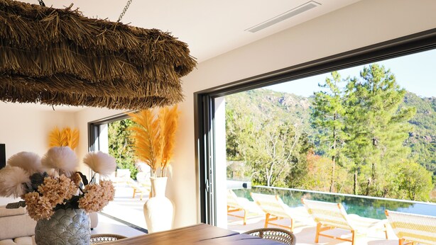 Un comedor moderno con una mesa de madera y sillas de mimbre, un jarrón decorativo con flores, y una vista a un paisaje montañoso a través de grandes ventanales.