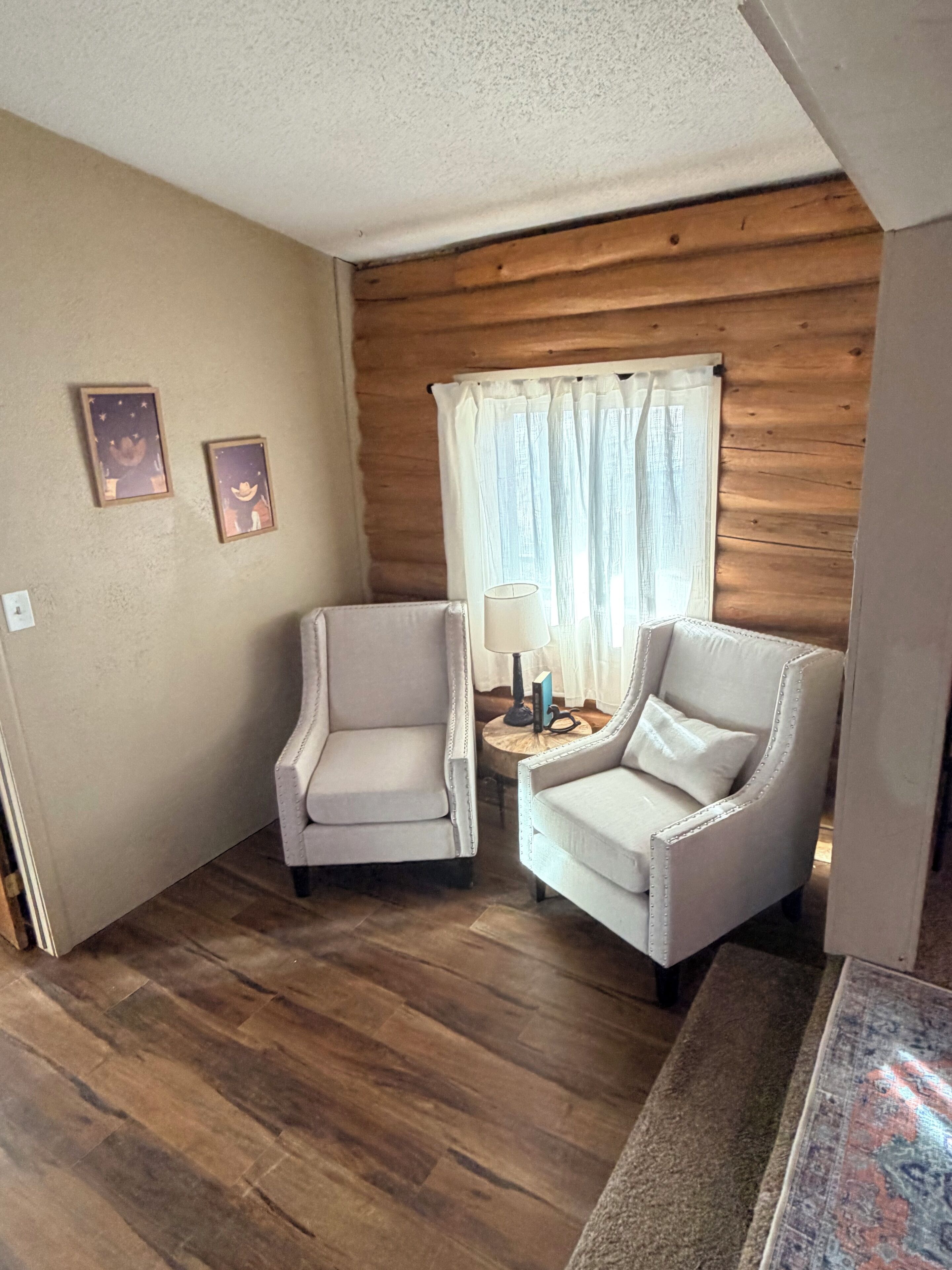 A room with two armchairs, a small round table with a lamp, and a window with sheer curtains.