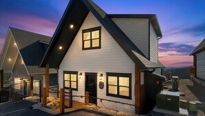 A modern two-story house with a steep gable roof, large windows, and a well-lit entrance. - New Lake-View Luxe Cabin w/ Pvt Hot Tub, 3/2 Sleeps 8! EV Charger, Branson, SDC! (Branson)