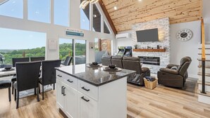 A modern kitchen with a stone backsplash, a flat-screen TV, and a dining area with black chairs and a white island. - New Lake-View Luxe Cabin w/ Pvt Hot Tub, 3/2 Sleeps 8! EV Charger, Branson, SDC! (Branson)