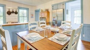 Un comedor con una mesa de madera para cuatro personas, sillas blancas y una cocina al fondo.