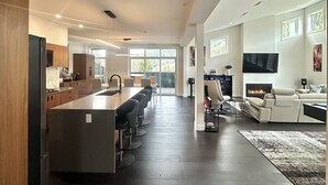Cozinha moderna e espaçosa, com ilha central, área de jantar com cadeiras, sala de estar com sofá e lareira, e uma TV fixada na parede.