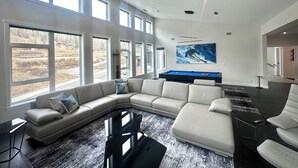 Uma sala de estar moderna com um amplo sofá em forma de L, mesa de bilhar e uma grande janela com vista para uma área residencial.