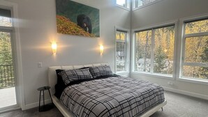 Quarto com uma cama grande, duas luminárias na parede, um quadro de um urso e grandes janelas com vista para árvores.