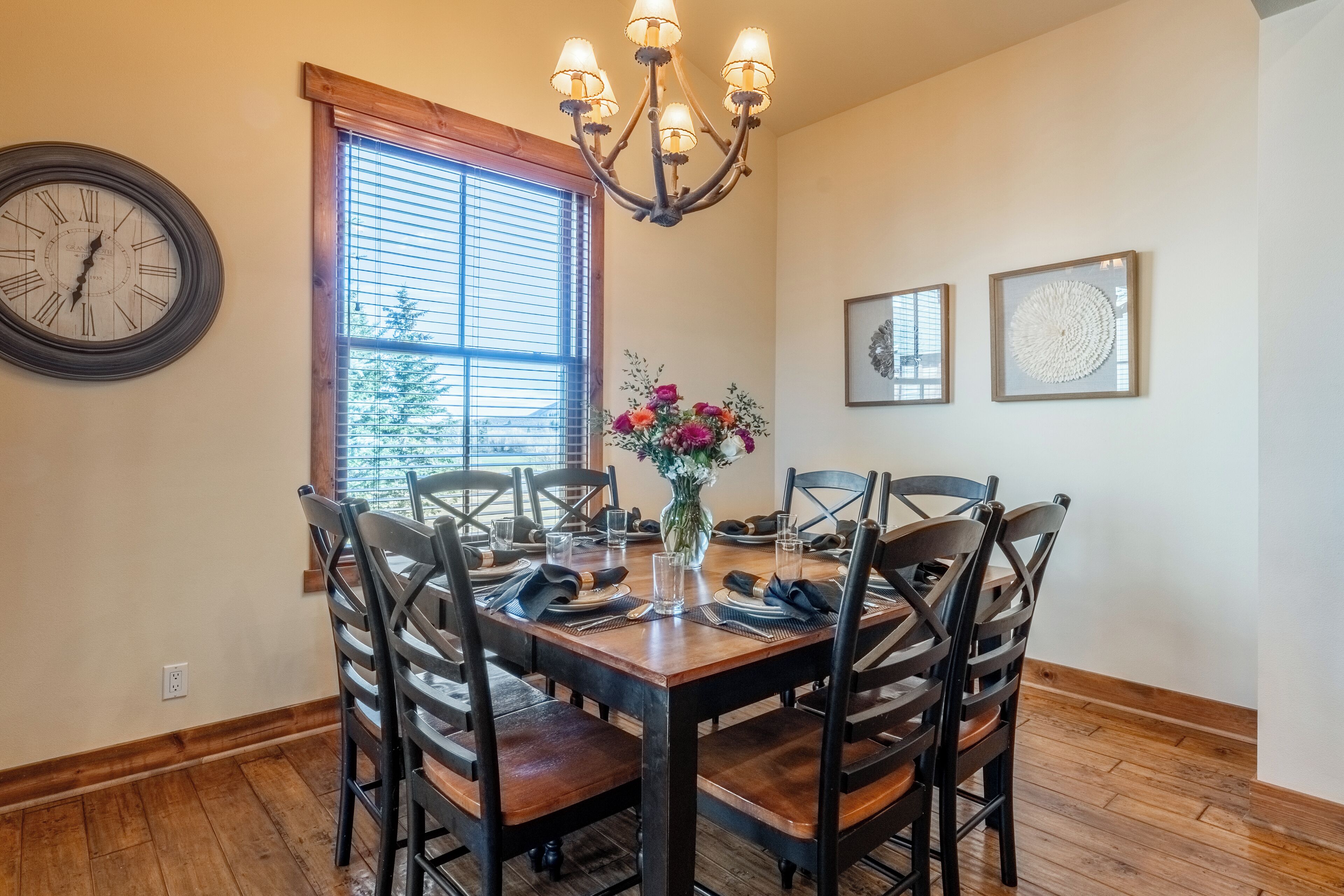 Un comedor con una mesa de madera dispuesta para cuatro personas, rodeada de sillas. Un candelabro cuelga sobre la mesa, y hay un reloj en la pared a la izquierda.