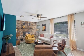 Un salón con pared de piedra, chimenea, ventilador de techo y un amplio ventanal con cortinas.