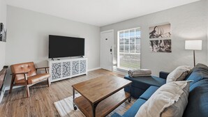 A living room with a flat-screen TV, a wooden coffee table, a blue sofa, a leather armchair, and a white cabinet with decorative panels. - Family, Business & Game-Day Getaway • Close to Arlington Attractions • Sleeps 8 (Arlington)