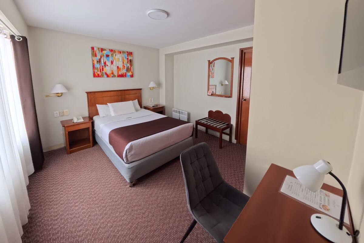 A hotel room with a bed, a desk with a chair, a small table, a lamp, a mirror, and a colorful abstract painting on the wall.