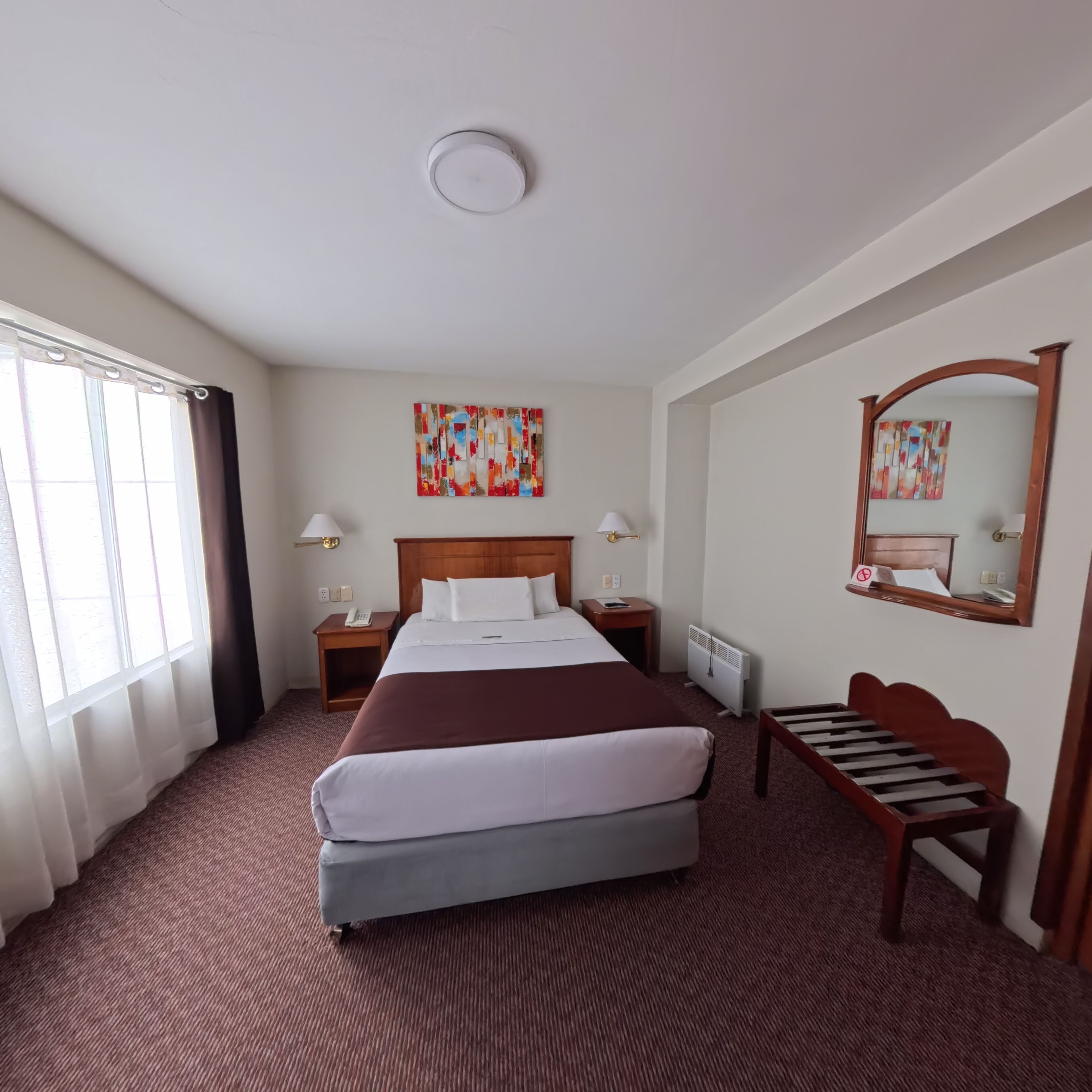 A hotel room with a large bed, a bedside table with a lamp, a radiator, and a wooden chair.