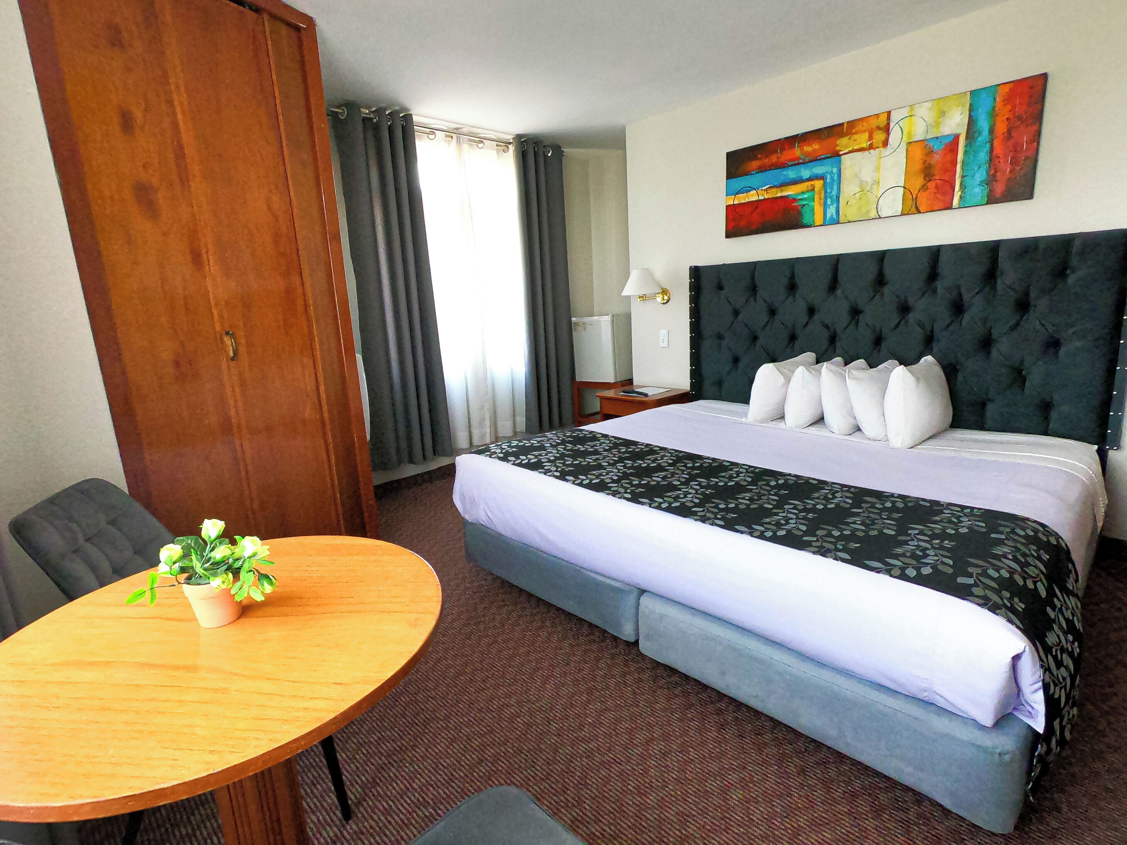 Ein Hotelzimmer mit einem großen Bett, einem Holzschrank, einem runden Tisch mit Pflanze und einem farbenfrohen abstrakten Gemälde an der Wand.