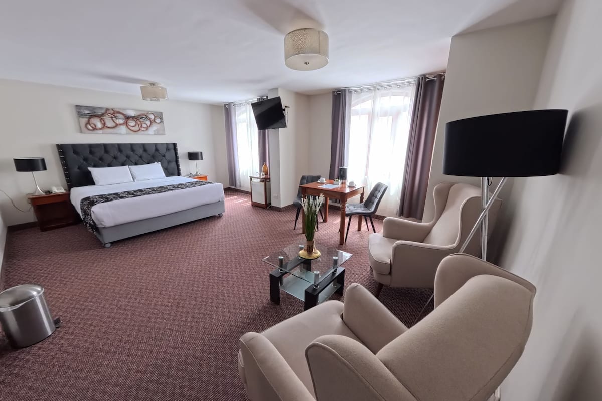 A hotel room with a bed, a desk, two chairs, a TV, and a large window with curtains.