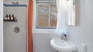 Un baño con ducha, lavamanos, ventana con vistas a un paisaje rocoso y un perchero con botellas.