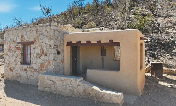A small stone building with a window and a door, surrounded by dry vegetation.
