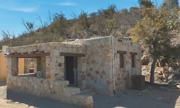 A small stone building with a door and windows, surrounded by trees and a clear sky.