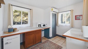 A compact kitchen with a refrigerator, microwave, and a small sink. There are two windows with curtains, and a rug on the floor.