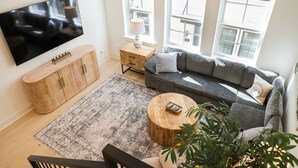 Un salon doté d’un grand téléviseur à écran plat, d’un meuble en bois, d’un canapé gris, d’une table basse ronde en bois et d’un tapis à motifs.