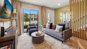 A modern living room with a grey sofa, a round coffee table, and a television. There is a large window with curtains, a painting on the wall, and a wooden partition. - Family townhouse w/ pool & workspace (Kissimmee)