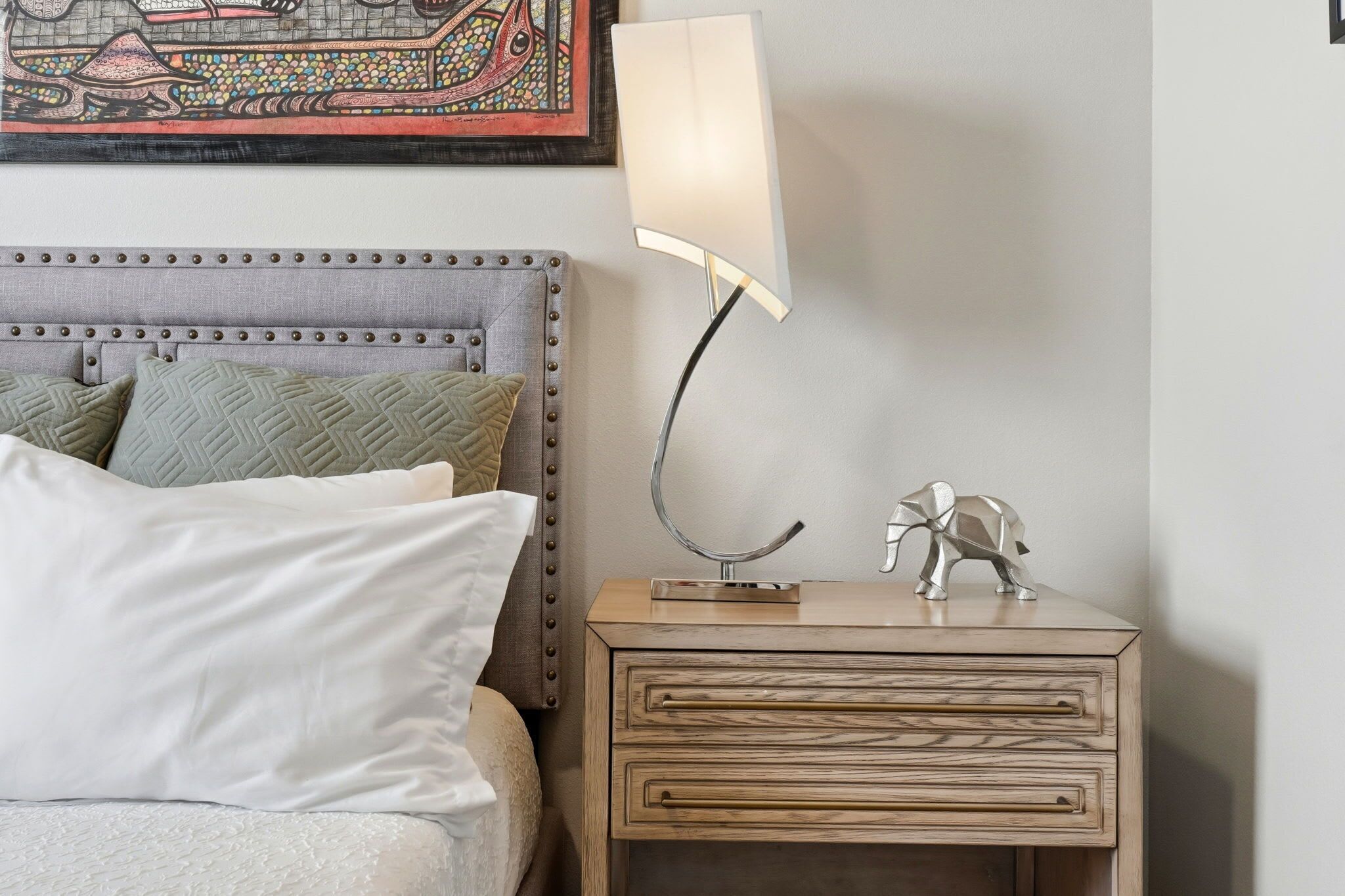 Un lit bien fait, avec une tête de lit, une lampe de chevet, une petite commode et une figurine d’éléphant décorative.