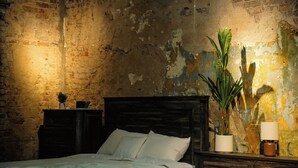 Een slaapkamer met een rustieke bakstenen muur, een houten hoofdbord, een plafondventilator en een nachtkastje met een plant en een lamp.