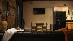 Een woonkamer met een zwarte leren bank, een kleine tafel met twee stoelen, een flatscreen televisie aan de muur en een keuken met houten kasten en een zwarte koelkast.